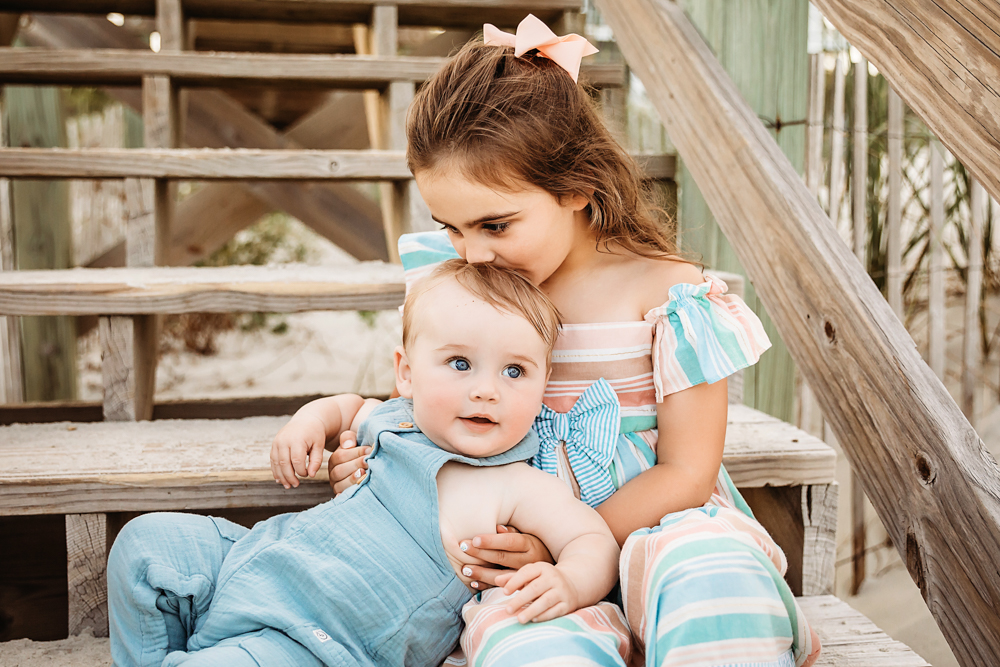 Stella kissing Coles head on weathered beach stairs, both in pastel outfits with soft sunset tones at Reflection Images by Tracy Rowell in Florence SC