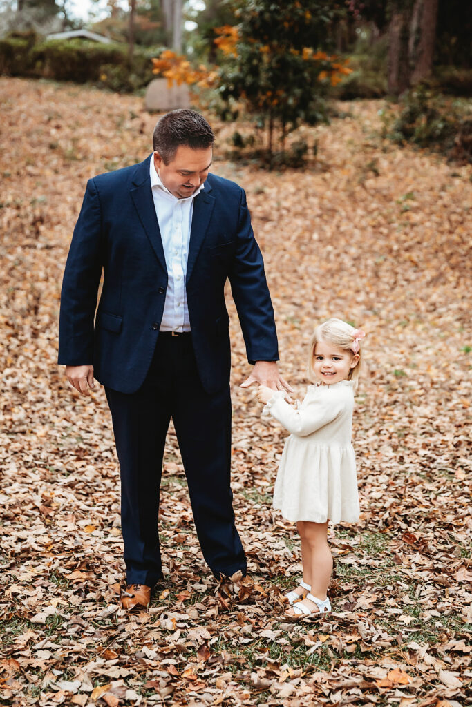 Father in navy suit standing with smiling daughter holding hands during fall family session at Reflection Images by Tracy Rowell