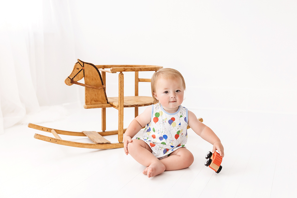 Levi sitting on a white floor with a small wooden toy in hand, smiling with a rocking horse behind him at Reflection Images by Tracy Rowell in Florence SC