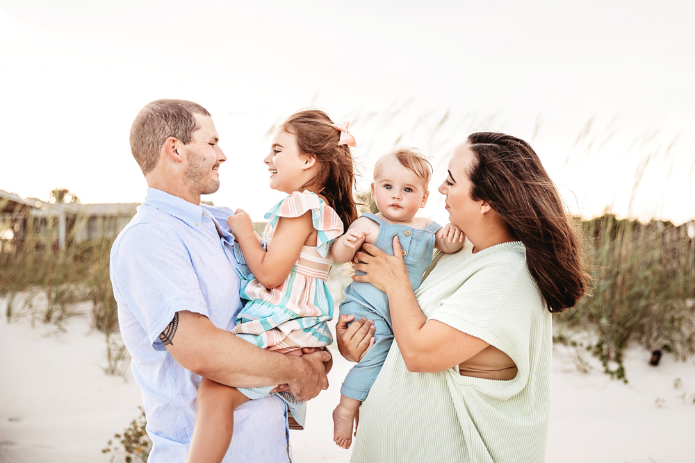 Skinner family laughing as Kristy and Chase hold Stella and Cole with windswept hair and white sand dunes at Reflection Images by Tracy Rowell in Florence SC