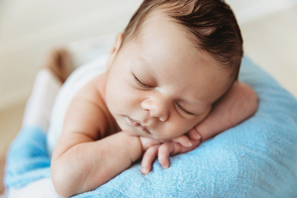 Top-down view of baby wrapped in blue, sleeping on arms with soft light and neutral studio background at Reflection Images by Tracy Rowell in Florence SC