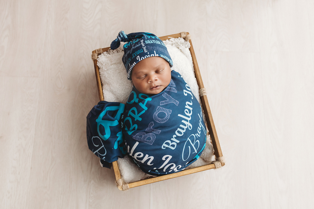 Braylen in custom blue name wrap and hat, sleeping in a box basket with white cushion on light wood floor at Reflection Images by Tracy Rowell in Florence SC