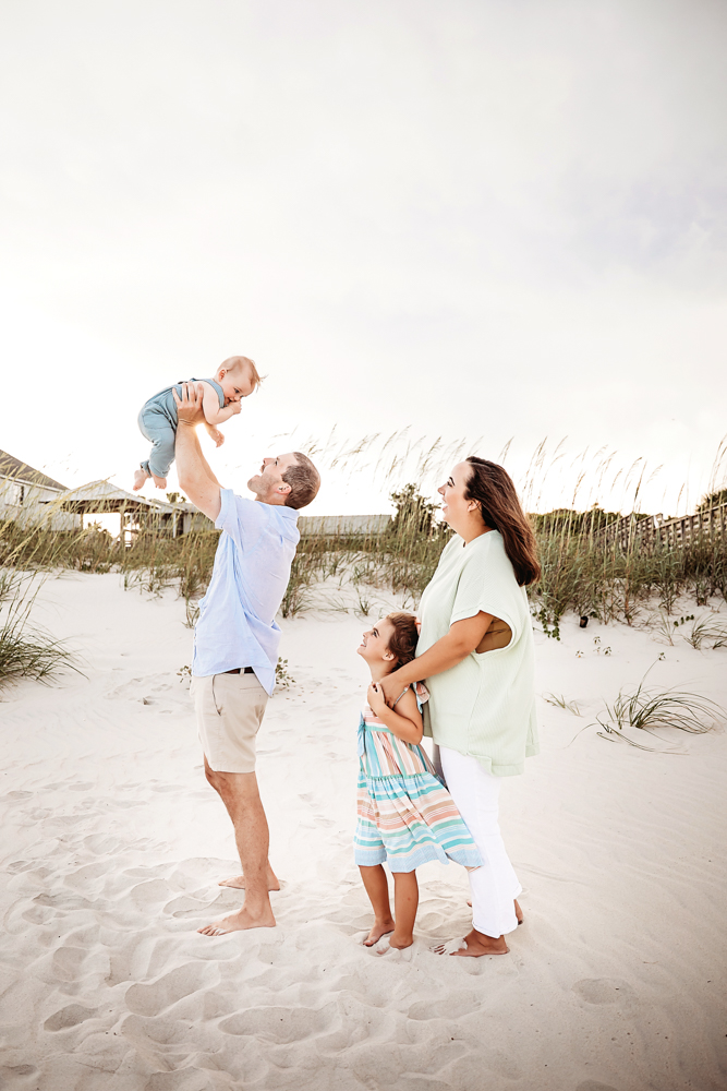 Chase lifting Cole high above with smiles from Kristy and Stella beside them on a sunlit beach at Reflection Images by Tracy Rowell in Florence SC