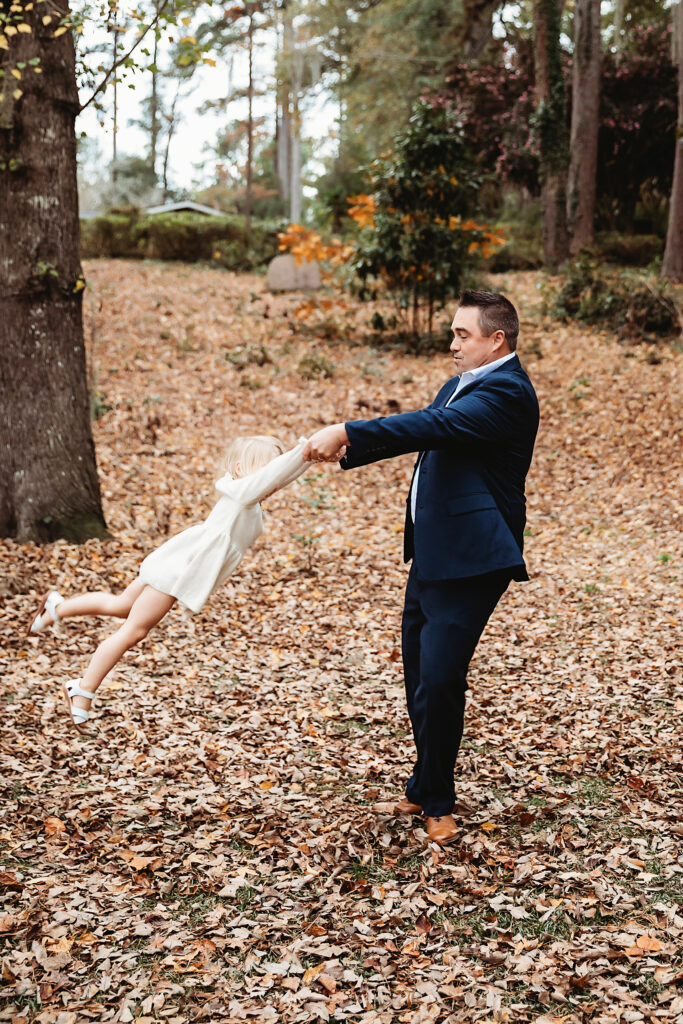 Playful dad spinning daughter in cream dress through autumn leaves in joyful moment captured in Florence SC