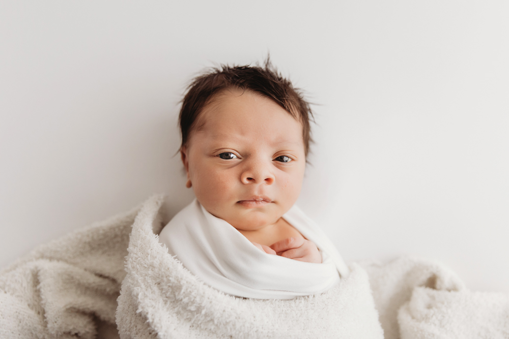 Awake portrait of newborn Liston swaddled in white, gazing gently while nestled in a cozy blanket at Reflection Images by Tracy Rowell in Florence SC