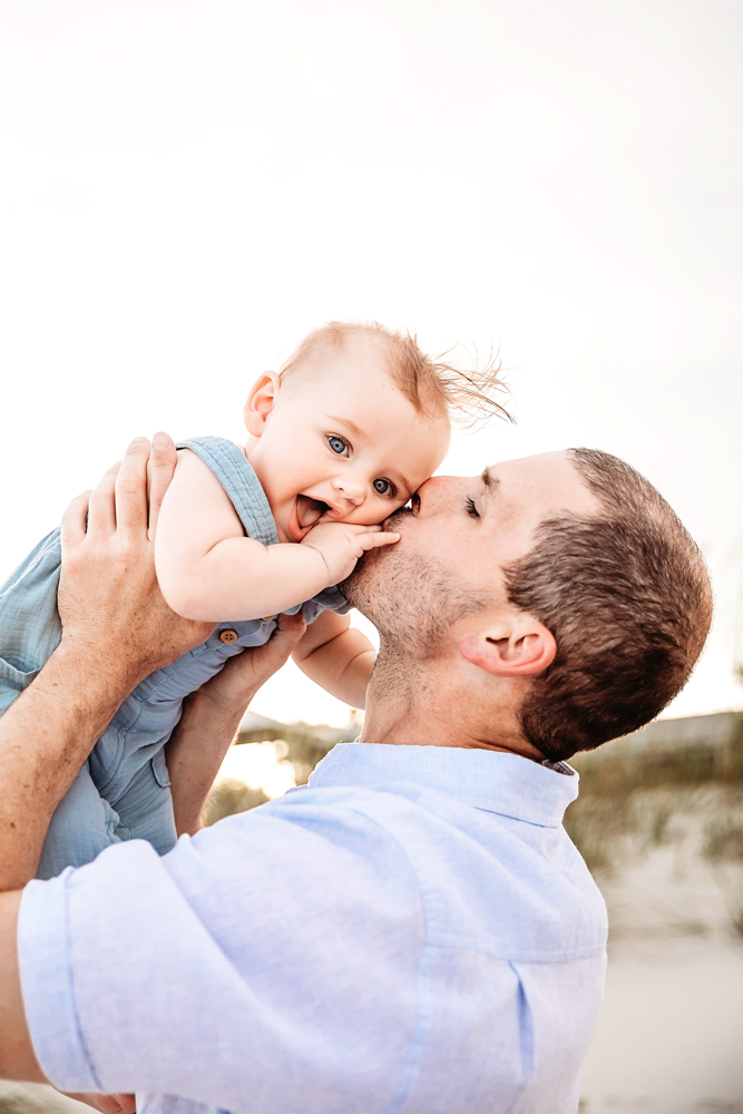 Cole being kissed on the cheek by dad as he giggles in the air with blue romper and bright sky at Reflection Images by Tracy Rowell in Florence SC