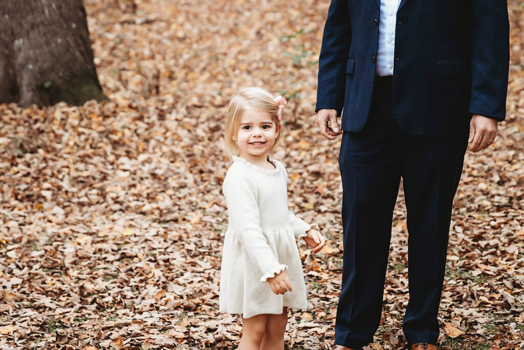 Little girl standing happily in front of dads legs during family photo shoot in forested park in Florence SC