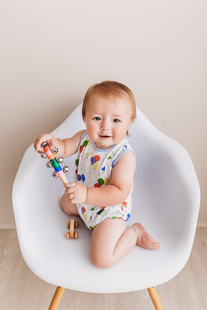 Levi sitting on a modern white chair holding a colorful tambourine and wooden car toy, playful and curious at Reflection Images by Tracy Rowell in Florence SC