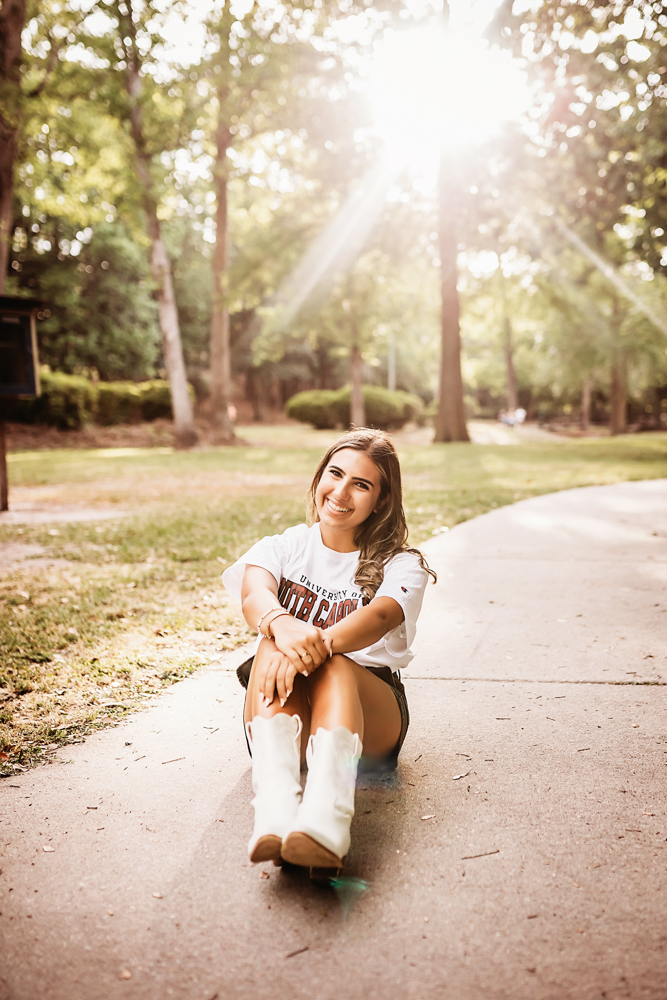 Senior session photo taken in wooded park at golden hour, showing a sitting pose in USC shirt and denim skirt from a close-up angle