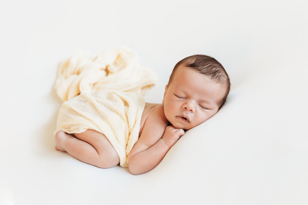 Newborn in pale yellow wrap laying sideways on white blanket with relaxed facial expression at Reflection Images by Tracy Rowell in Florence SC