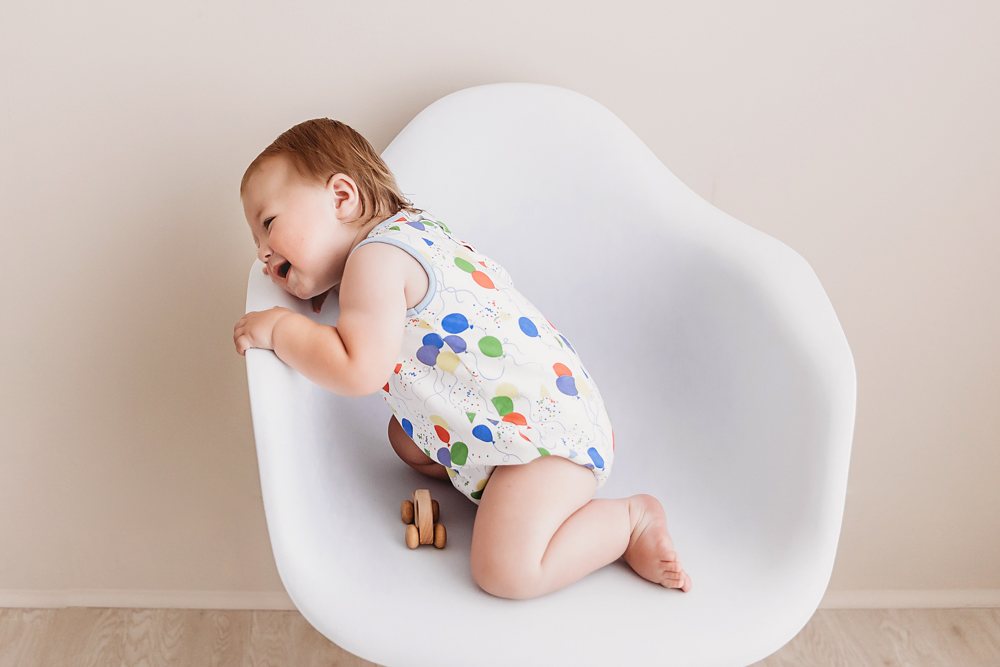 Levi leaning joyfully across a white chair in balloon romper with wooden toy nearby on light wood flooring at Reflection Images by Tracy Rowell in Florence SC