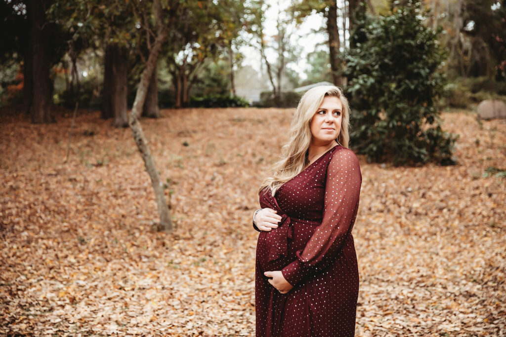 Expecting mother in long burgundy gown looking off to the side while cradling belly, surrounded by fall foliage in Florence SC