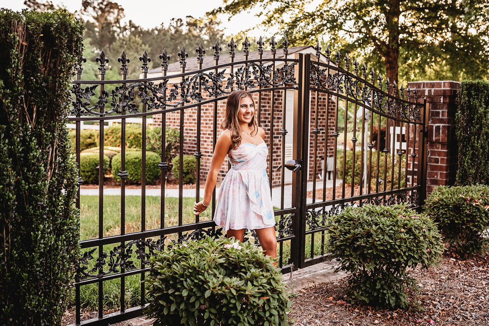 Senior session photo taken in sunlit trail at golden hour, showing a smiling pose in floral dress from a full-body angle