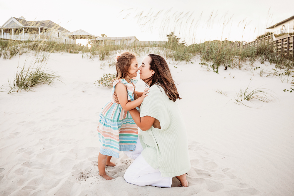 Kristy kneeling in the sand to kiss Stella on the forehead with dune grass and beach cottages behind at Reflection Images by Tracy Rowell in Florence SC