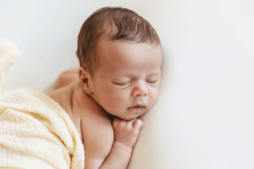 Close view of baby sleeping on white surface with soft yellow wrap and tightly curled hands near face at Reflection Images by Tracy Rowell in Florence SC