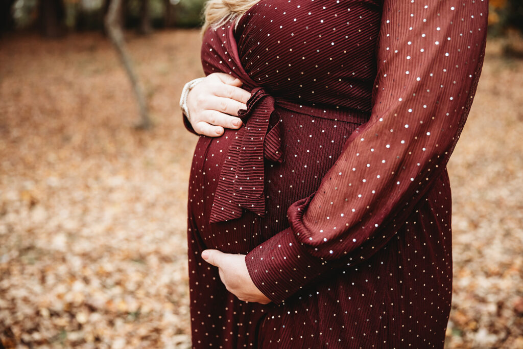 Close-up of pregnant belly with hands cradling bump, mother wearing burgundy polka dot dress during autumn maternity shoot