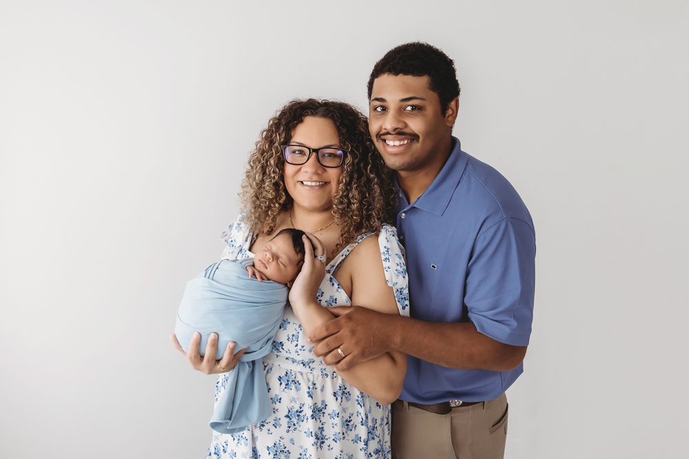 Smiling family portrait of mom and dad holding newborn Liston in blue wrap on white background at Reflection Images by Tracy Rowell in Florence SC