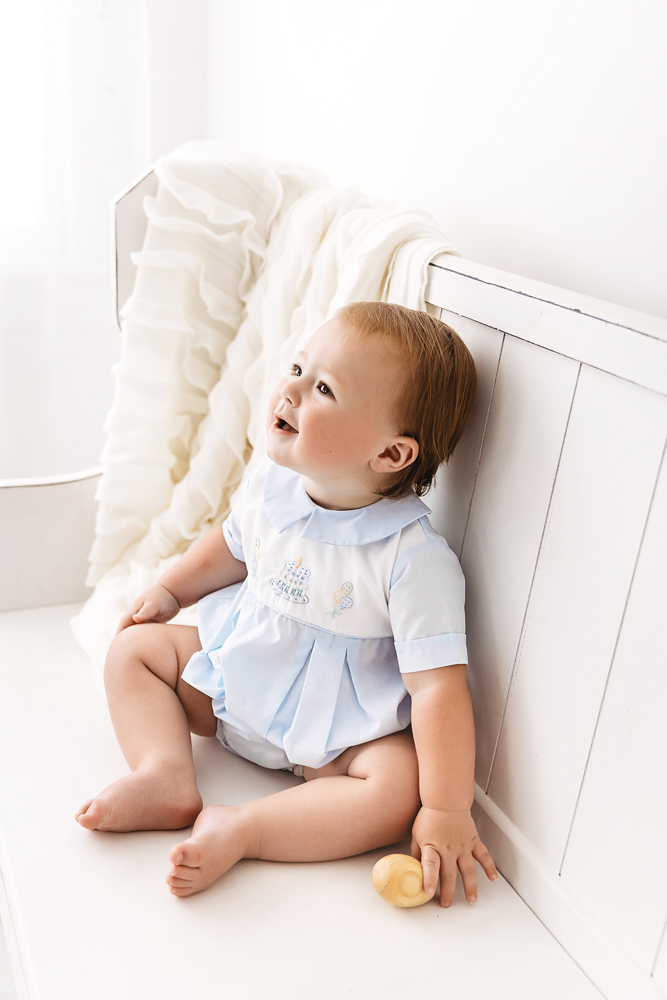 Levi seated on a white bench with a cream ruffle blanket, holding a wooden toy while smiling upward in a pale blue romper at Reflection Images by Tracy Rowell in Florence SC