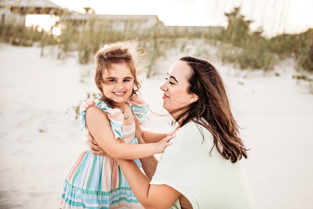 Stella grinning while being hugged by Kristy on the sunlit beach with windswept grass and pastel tones at Reflection Images by Tracy Rowell in Florence SC