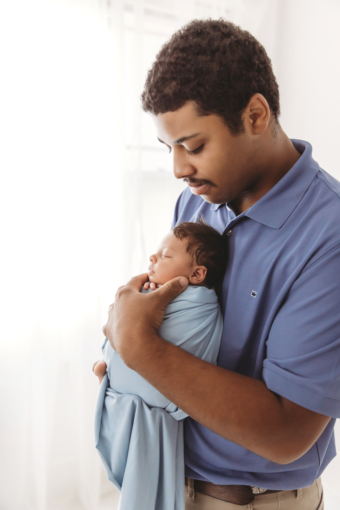Dad holding sleeping newborn Liston close, wrapped in blue, softly lit by natural window light at Reflection Images by Tracy Rowell in Florence SC