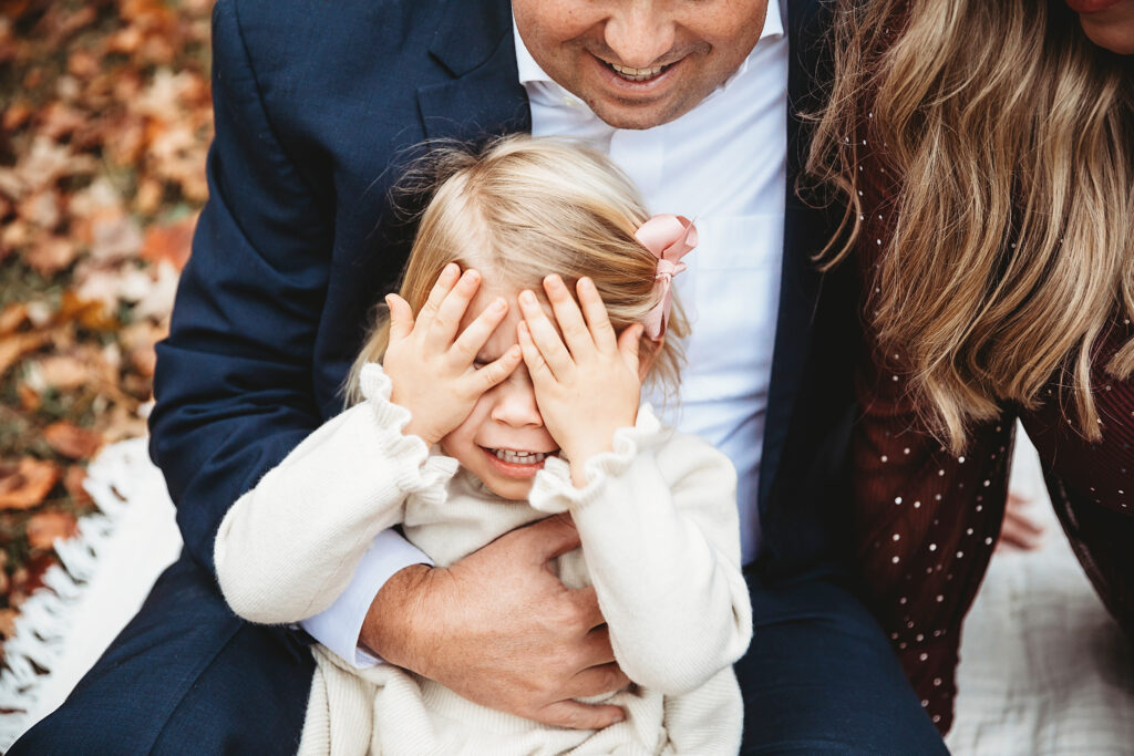 Daughter playing peekaboo with hands over face while sitting on dads lap during playful family session at Reflection Images