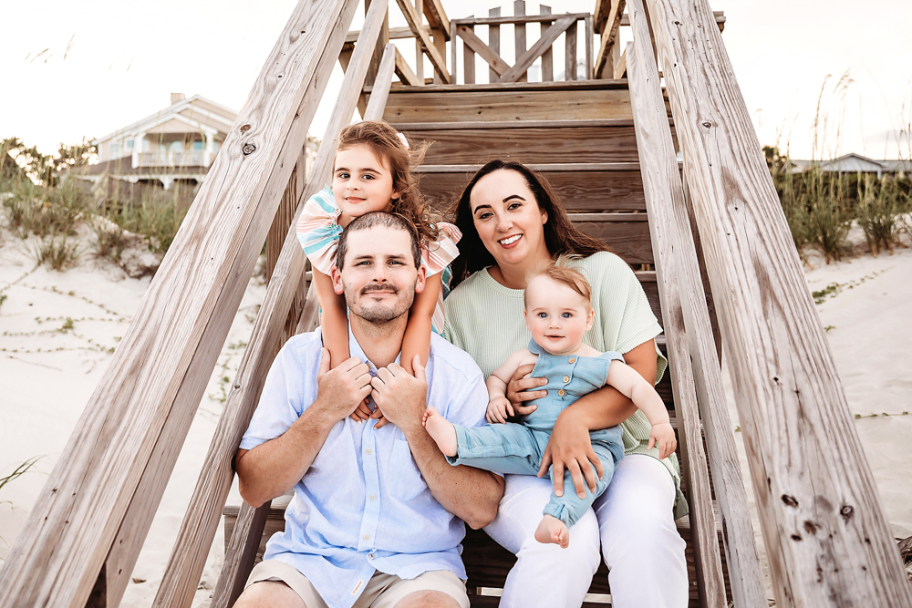 The Skinner family posed on wooden beach stairs with Stella on Chases shoulders and Cole in Kristys arms at Reflection Images by Tracy Rowell in Florence SC