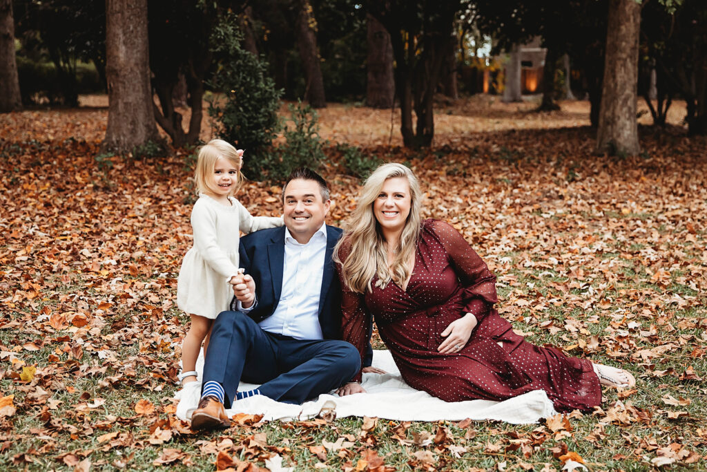 Happy family seated on blanket, mom and dad smiling as daughter stands between them in charming autumn portrait