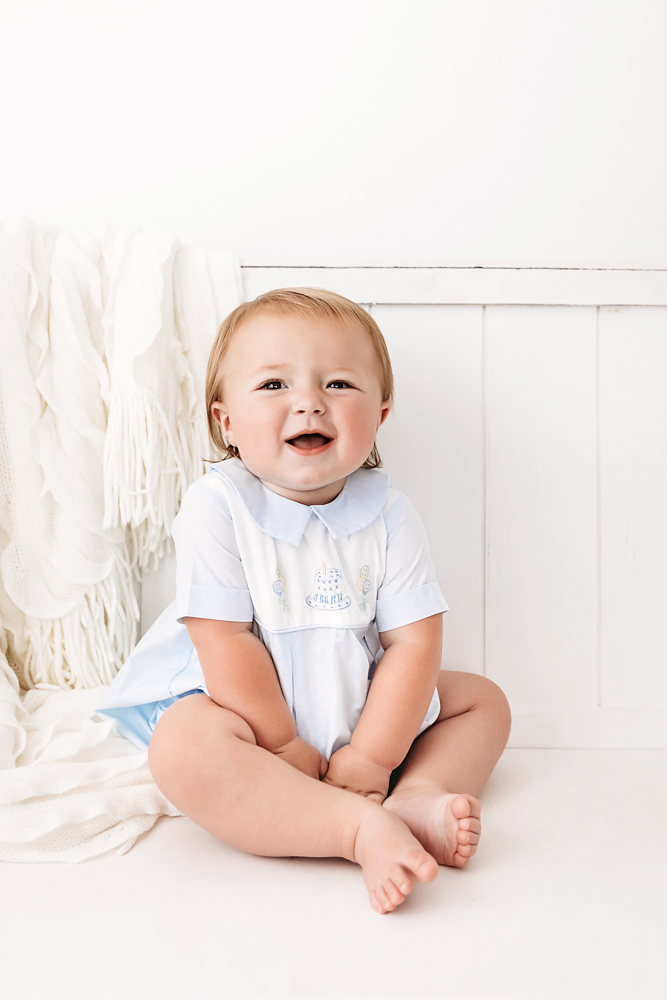Levi smiling brightly while sitting cross-legged in front of a white wall and soft knit blanket, wearing a classic romper at Reflection Images by Tracy Rowell in Florence SC
