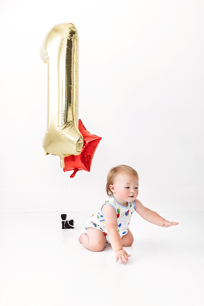 Levi crawling toward the camera with a large gold '1' balloon in the background, wearing a colorful romper on a white studio floor at Reflection Images by Tracy Rowell in Florence SC