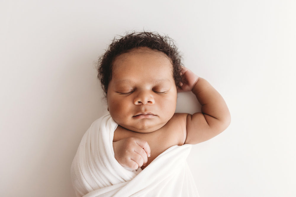 Braylen peacefully sleeping with one arm raised and wrapped in soft white swaddle on a white backdrop at Reflection Images by Tracy Rowell in Florence SC