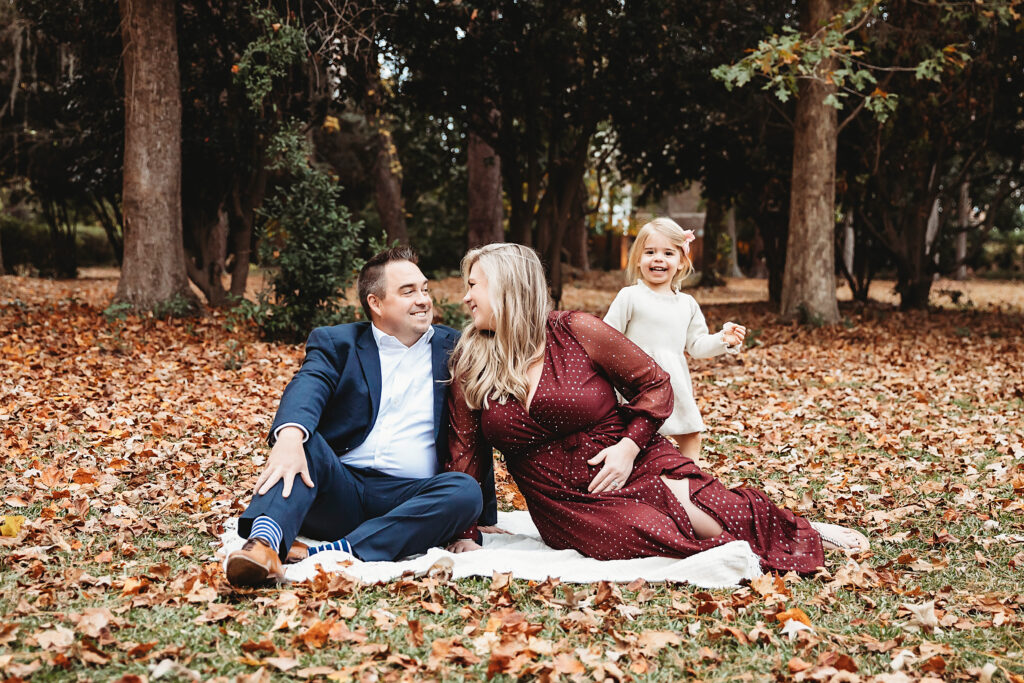 Family smiling on blanket during maternity session with daughter standing behind mom and dad among fallen leaves in Florence SC