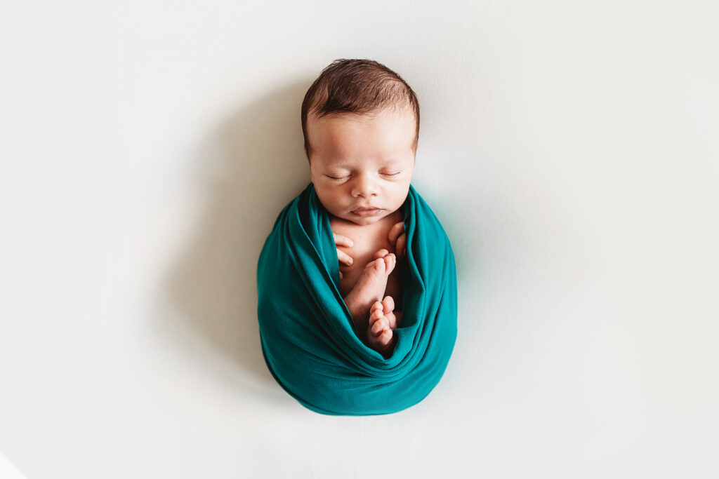 Full view of newborn wrapped snugly in teal swaddle with feet peeking out, centered on white backdrop at Reflection Images by Tracy Rowell in Florence SC