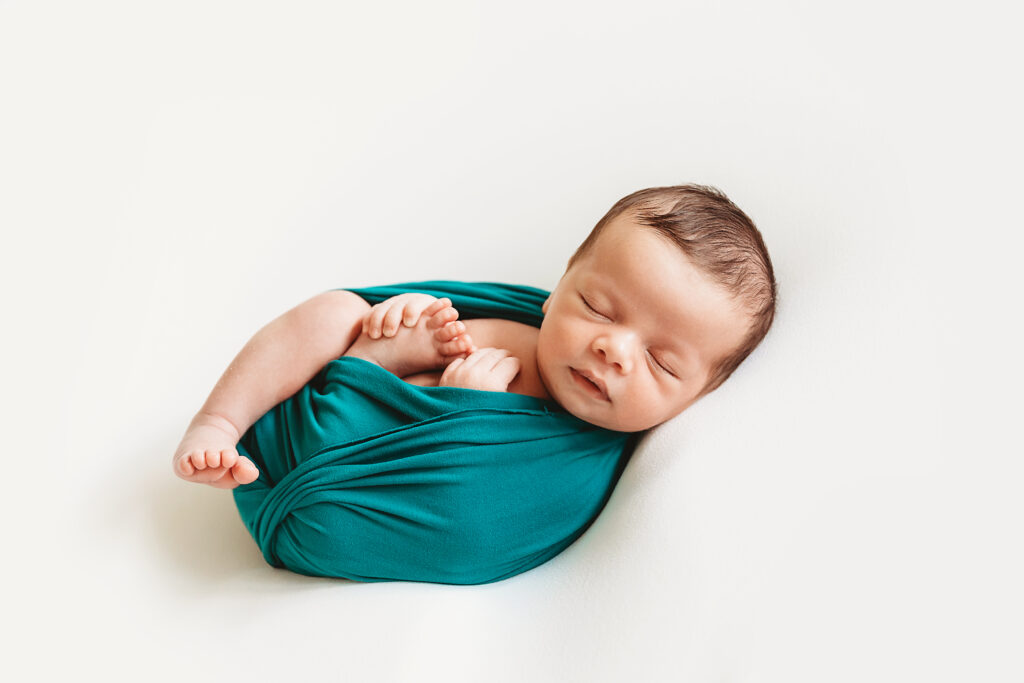 Newborn baby smiling while wrapped in green cloth, lying on a white background
