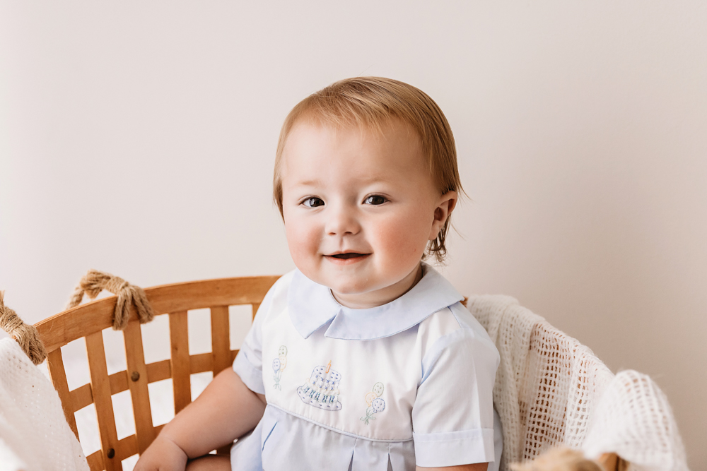 Baby Levi sitting in a wicker basket with a joyful expression, wearing a light blue romper with embroidered balloons, on a white flokati rug at Reflection Images by Tracy Rowell in Florence SC.