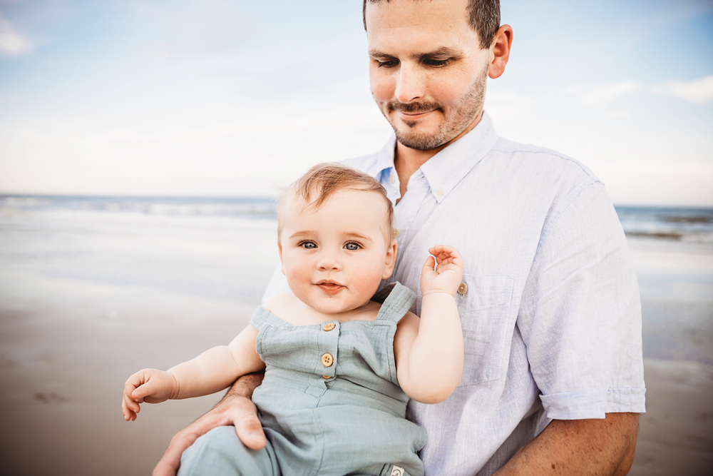 Close-up of Cole in a pale blue romper held in Chases arms with gentle beach light and ocean backdrop at Reflection Images by Tracy Rowell in Florence SC