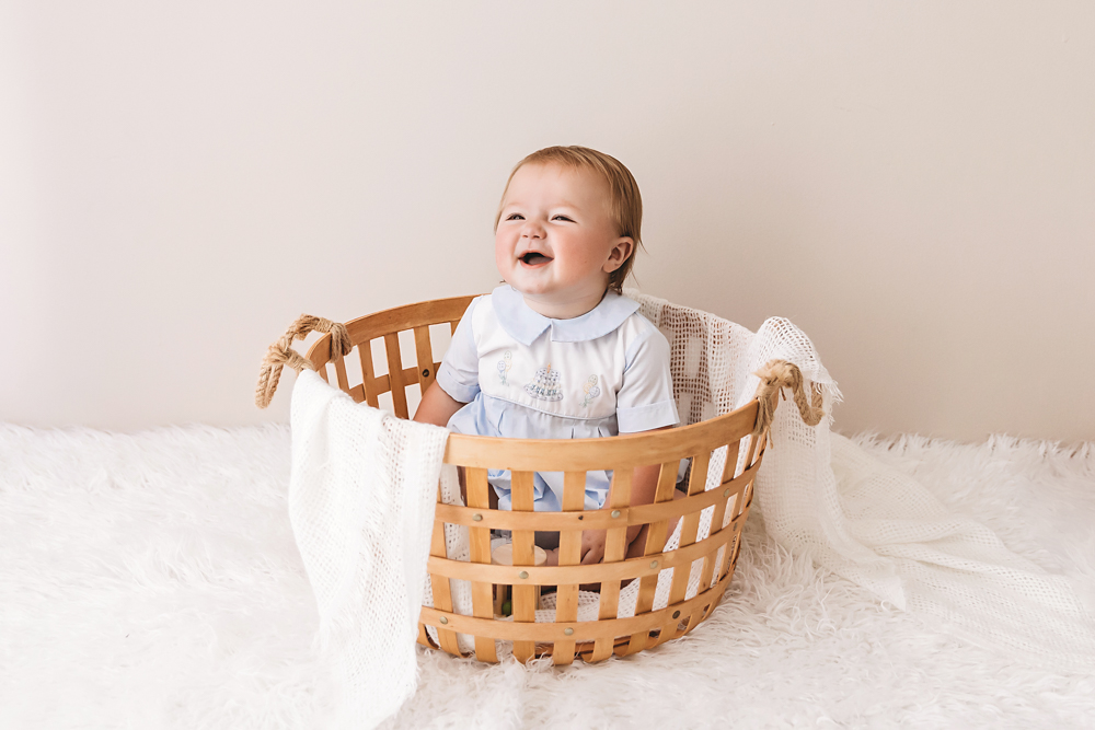Close-up of Levi smiling in a woven basket, wearing a classic blue birthday romper against a neutral backdrop at Reflection Images by Tracy Rowell in Florence SC.