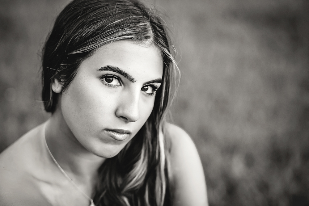 Black and white Senior session photo taken in sunlit trail at golden hour, showing a sitting pose in white boots from a over-the-shoulder angle