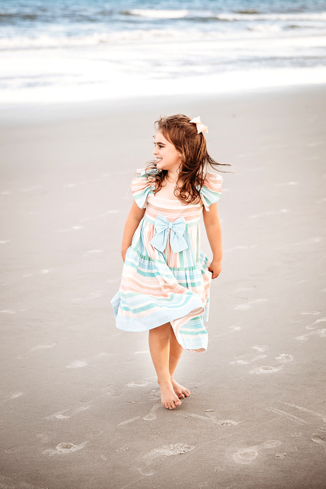 Stella walking barefoot along the shore in a pastel-striped dress with bow, soft waves in background at Reflection Images by Tracy Rowell in Florence SC