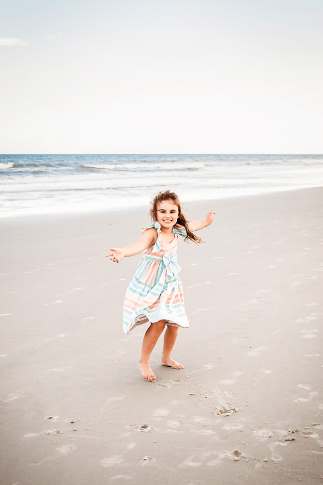Stella running toward the camera with open arms and a wide smile, her striped dress flowing on the beach at Reflection Images by Tracy Rowell in Florence SC