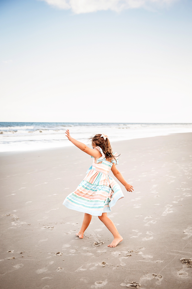 Stella mid-twirl in a striped dress, arms raised as wind catches her curls on the sand at Reflection Images by Tracy Rowell in Florence SC