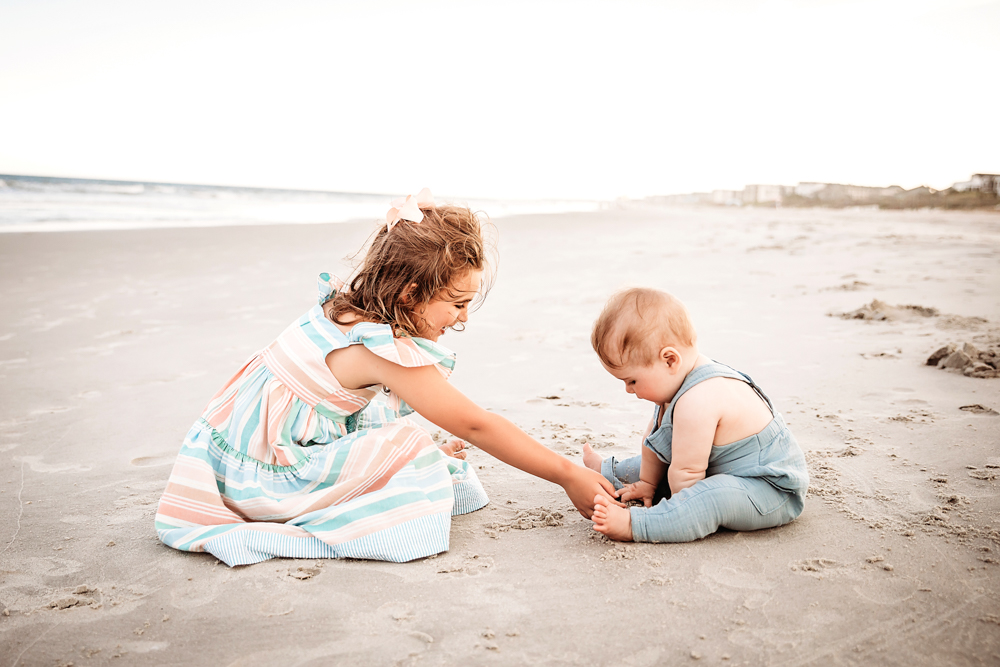 Stella gently reaching for Coles feet as they sit barefoot in the sand, ocean and soft glow behind at Reflection Images by Tracy Rowell in Florence SC