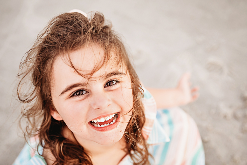 Close-up of Stellas joyful expression with wind-blown curls and soft pastel dress in beach light at Reflection Images by Tracy Rowell in Florence SC