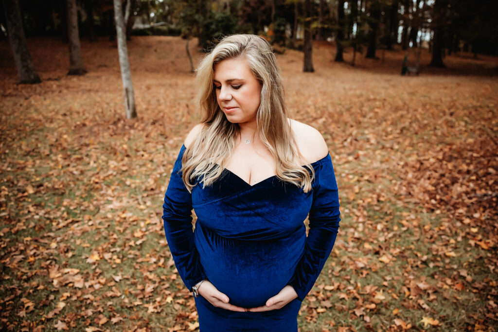 Confident pregnant woman smiling in blue velvet gown surrounded by orange autumn leaves in Florence SC