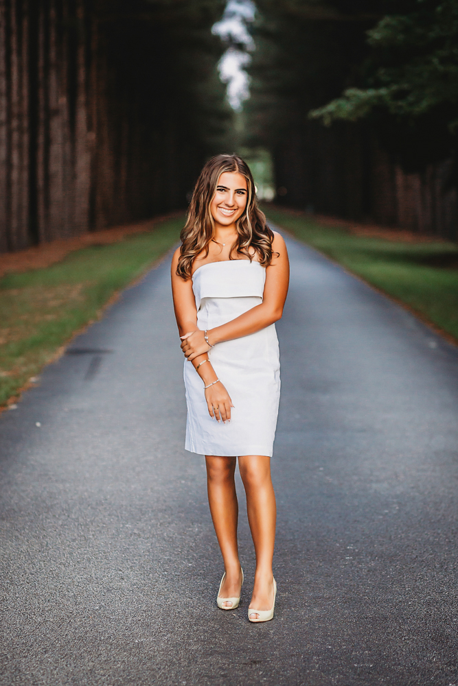 Senior session photo taken in wooded park at golden hour, showing a walking pose in white dress from a over-the-shoulder angle