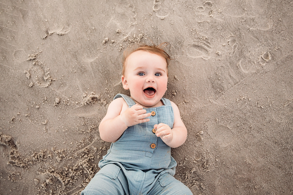 Top-down view of Cole giggling on the sand in a light blue romper with tousled hair and button detail at Reflection Images by Tracy Rowell in Florence SC