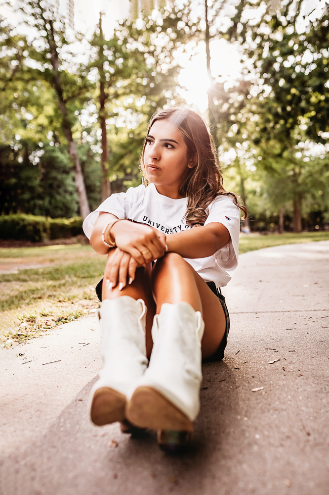 Senior session photo taken in gravel path at golden hour, showing a sitting pose in casual style from a eye-level angle