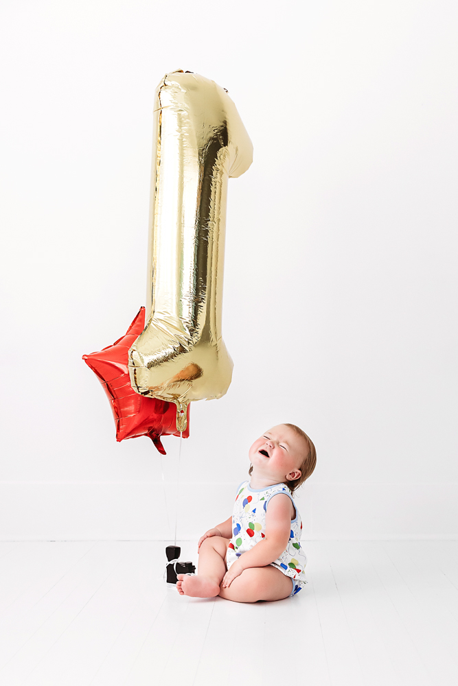 Levi smiling as he sits next to a gold number 1 balloon and red star, captured on a white floor during his birthday session at Reflection Images by Tracy Rowell in Florence SC