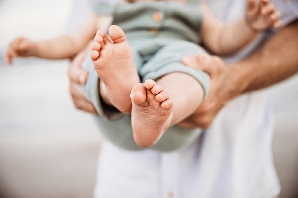 Close-up of Coles tiny feet held forward in Chases hands, soft focus on beach tones behind at Reflection Images by Tracy Rowell in Florence SC