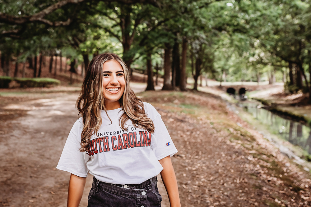 Senior session photo taken in rustic bridge at golden hour, showing a smiling pose in USC shirt and denim skirt from a close-up angle