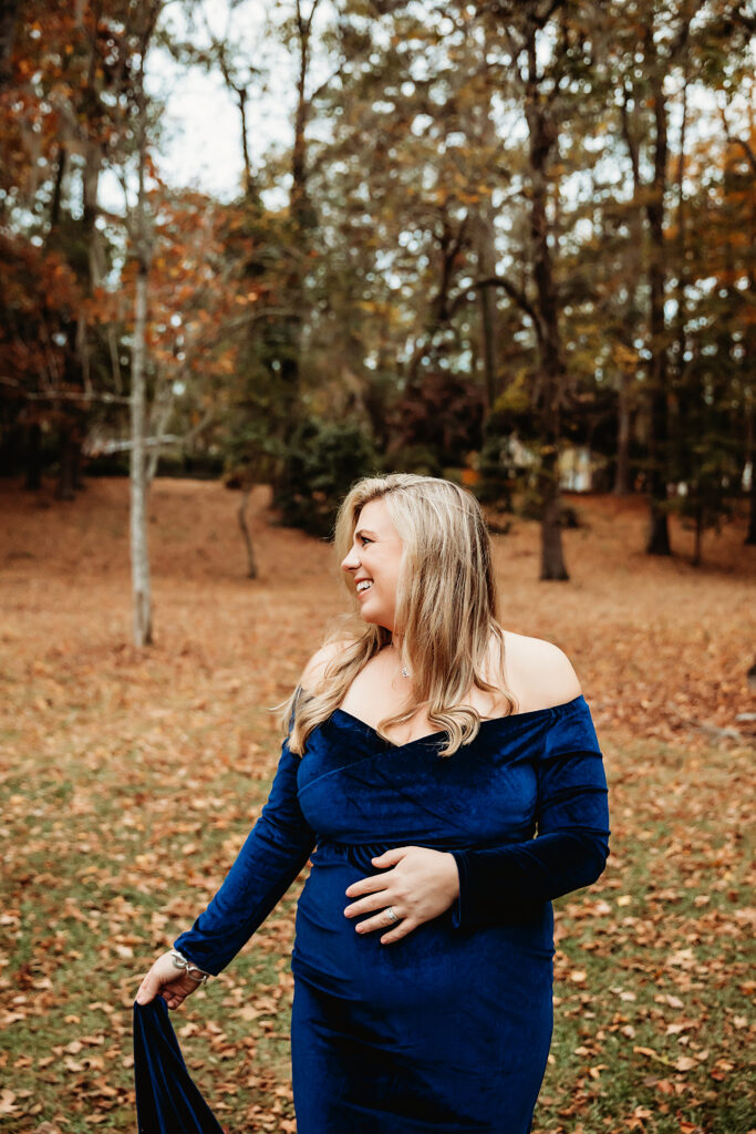 Expecting mom in blue velvet gown smiling joyfully in autumn park with leaves on the ground in Florence SC